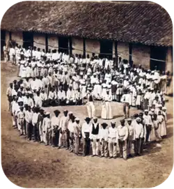 Photograph showing a group of people dressed in white, who have gathered in front of a tile-roofed farm building and observe another large group which has formed a large circle surrounding five men straddling large drums, a woman and two other men