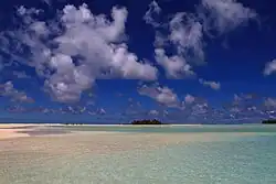 Very shallow water over a white seafloor with a green island in the background beneath blue sky and scattered clouds