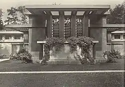 Low building with ivy draped terrace and three prominent vertical windows with tall trees in the background