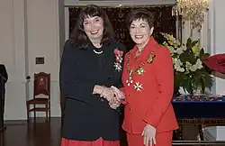 Two smiling women face the camera shaking hands. They are wearing formal clothes.