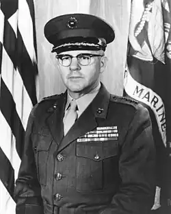A stocky Caucasian man with brown hair and thick glasses in military uniform in front of a flag