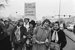 A black and white image of demonstrators marching with linked arms and carrying signs in support of Eliane Morissens's hunger strike.