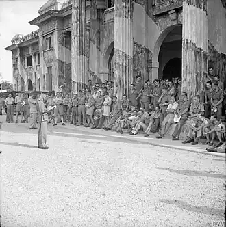 A large group of officers stand in front of a colonnaded building; a senior officer addresses them.