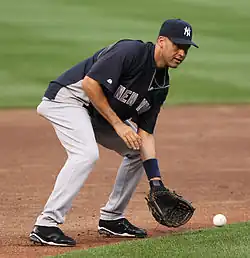A man in a blue baseball uniform with "New York" written on the front in gray letters and a navy hat with white letters "N" and "Y" interlocking prepares to catch a ground ball with his baseball glove.