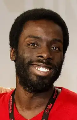 close-up photo of Desmond Cole wearing a bright red t-shirt with a black lanyard, grinning directly at camera