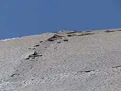 An inclined wall covered with white pieces of ceramic under a blue sky. Some of the tiles are missing, others are detached from the wall and raise against other pieces.