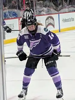 Photo of Dom Petrie, a redheaded woman in a purple Minnesota Frost hockey jersey, standing with with her stick on her knees before a faceoff.