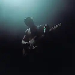 The image depicts Ross Lynch at the center, playing guitar on a dark greenish background and a low light illuminating him.