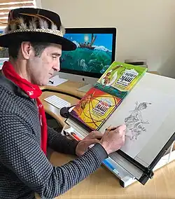 A man in a top hat decorated with feathers sketches a drawing of a young Māori boy, next to copies of his books. A finished illustration is seen on a computer in the background, showing an ocean scene from the legend of How Māui Fished Up The North Island.