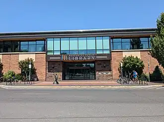 Photograph of the Downtown Bend Library, a modern brick and glass building, on a sunny day. Above the doors is a sign that reads Deschutes Public Library and a minimalist logo of multicolored vertical lines representing books. A metal statue appears in front of the library. There are bike racks on either side of the entrance. Two people approach the library afoot.