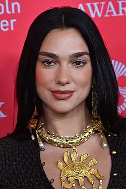 Headshot of Dua Lipa, a young, white woman with long, dark hair wearing a shimmery silver outfit
