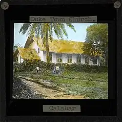 Image of the Duketown Church, Calabar (located within later day Nigeria). Three people stand in front of the white-sided church with a thatched roof. Duketown lies on the Calabar River 50 miles from the coast.