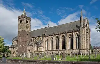 Side view of the nave of a cathedral from outside. Tall arched glass windows run along half the length of the nave from the right. Adjacent to the nave, and the left of the scene is a cuboid-shaped tower with a conical spire. The foreground is scattered with headstones of a graveyard on green grass.