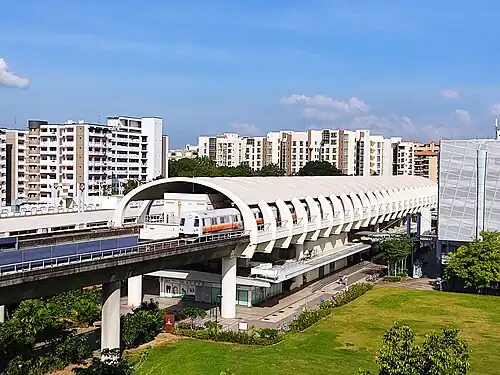 A bird's eye shot of the station, which is a white caterpillar-like segmented dome in the centre. There are a variety of buildings next to the station. A train is also coming out of the station