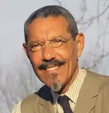 a tan skinned man with narrow glasses smiling in a tight headshot in an sunny outdoor photo