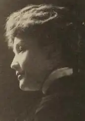 A white woman in profile, brightly lit from the left; her hair is in an updo, and she is wearing a high-collared blouse and jacket.