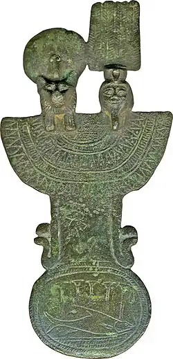 This is a menat counterweight made of bronze. It depicts the heads of Shu (rightmost head) alongside his sister Tefnut, goddess of moisture (leftmost head). Tefnut is rendered as a lioness with a mane wearing a uraeus, wig, and sun disk. Shu is shown dressed in a false beard, shorter wig, and a crown that includes a modius, tall feathers, and a uraeus. Two serpents flank the counterweight near the bottom where an oxyrhynchus fish can also be seen.