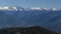 A range of snow-capped mountains. In the foreground is a stretch of forest. In between is an area of lower ground.