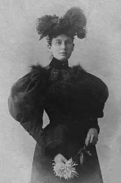 In this three-quarter length portrait, Ethel Ogden dressed in a black puff-sleeved dress with a high collar looks directly at the viewer. She is wearing a hat adorned with feathers and holds a long-stemmed spider mum flower in her hands.