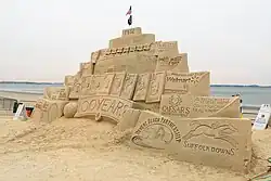Pyramid shaped sand sculpture featuring Fenway Park's green monster wall at the center and various sponsors of the event on either side. The sculpture says 1912, with an American flag and POW flag sticking out the very top. Below that are the words Fenway Park and details of the green monster, and a collection of presumably former Red Sox players represented by baseball cards lined up above a banner reading "100 years." Along the edges of the pyramid, sponsors including Honey Dew, Walmart, Massachusetts General Hospital, and Suffolk Downs are visible.
