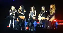 Five young women wearing black clothing sitting on tall chairs in a line on a stage.