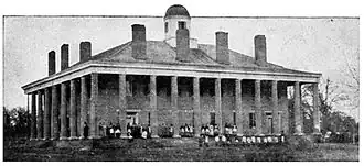 Two-story Greek revival style building with a colonnade surrounding the porch, seven chimneys visible on the roof, and a cuppola at its peak. A group of women are standing in the yard.