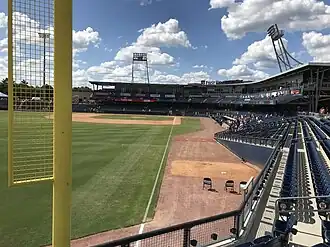 A green baseball field surrounded by its brown shale warning track and blue seats viewed from the concourse by the yellow left field foul pole on a sunny afternoon