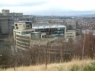 A modern office building featuring beige stone walls, large glass windows, and multiple terraces with greenery. The photo is taken from an adjacent hill, and leafless trees are visible in the foreground.