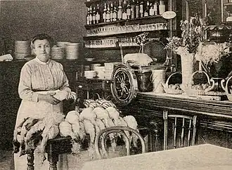white woman in late 19th-century clothes photographed in a kitchen, with a pile of chickens on the table