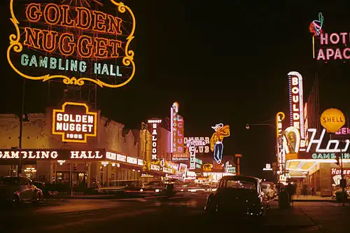 An old photograph of a busy street at night. There are large neon signs on every building.