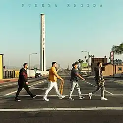 Four men walking on a sidewalk, with the Santa Fe Smokestack and a hotel seen in the background.