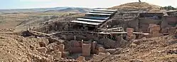 Roughly built stone walls surrounding T-shaped stone pillars under a modern steel walkway and roof in a hilly landscape