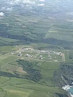 A view of GCHQ Bude from the air, taken from a small aircraft from a couple of miles away. The Atlantic Ocean is visible in the bottom right of the image and GCHQ Bude itself lies on a cliff face amongst rolling fields