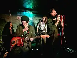 First man looks towards his left, he wears a dark jacket over white tee-shirt. Second man looks down and to his right, only his left hand holds the guitar's neck, while his right hand is up near his left shoulder. Third man has his head twisted to his left while playing his bass guitar. Fourth man, 30 years-old, holds his microphone with both hands as he sings. Behind the performers a partly obscured sign reads "Banquet Records"