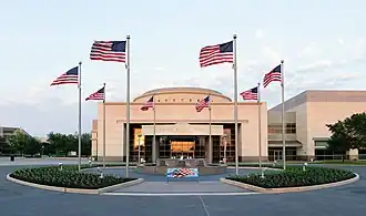 Above the doorway of a large, relatively plain rectangular structure with a short dome are the words "George Bush Library". In front of the building is a circular courtyard with a water fountain; eight American flags are positioned evenly around the circle.