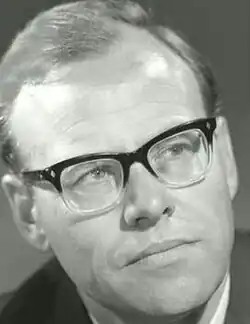 Publicity headshot of clean-shaven man with pale, oiled hair, brushed back, and spectacles.