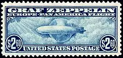A rectangular postage stamp, torn from a perforated sheet. It is in landscape format and is printed with blue ink on a white background. At the top it says "Graf Zeppelin: Pan-American Flight" and on the bottom "$2.60 United States Postage $2.60". In between is a depiction of the airship, flying from right to left, against a background of the planet Earth. Stylised clouds swim across the scene, and rays of sunlight are shining in from the top-left corner.