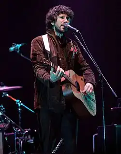 A man with long, bushy dark brown hair and a short beard playing acoustic guitar while singing into a microphone and looking to his right, away from the camera. He is seen from the knees upwards and is wearing a brown jacket with a red and blue embroidered pattern on the right shoulder.