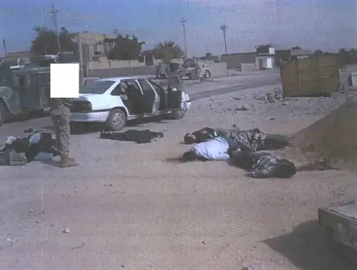 The bodies of five Iraqi civilians are laid out on the ground by the car they were shot in, as a Marine stands over them.