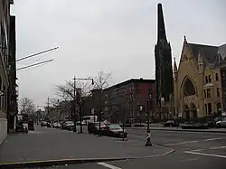 A two-lane street passes through a city neighborhood composed of a line of three-story buildings and a large church. The street is lined with decorative lampposts and small trees.