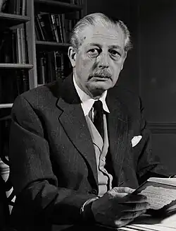Harold Macmillan, wearing a jacket and tie, sitting at a desk