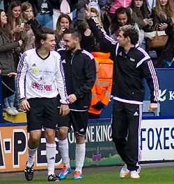 Three young adult men, Harry Styles, Liam Payne, and Niall Horan, seen walking on a football field together wearing Adidas tracksuits. Payne and Horan face the left of the image and wave towards the stadium filled with fans, whilst Styles looks the other way.