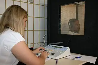 a female medical professional is seated in front of a special sound-proof booth with a glass window, controlling diagnostic test equipment. Inside the booth a middle aged man can be seen wearing headphones and is looking straight ahead of himself, not at the audiologist, and appears to be concentrating on hearing something