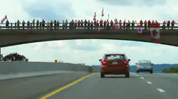 A bridge showcased against the sky, with the ground not visible. Lining the bridge are people, some holding Canadian flags.