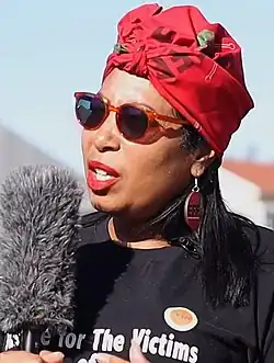 A woman holding a microphone, wearing a red headscarf, sunglasses, large solid earrings with "EFF" inscribed, and a black t-shirt
