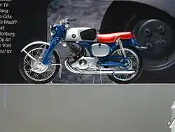 A pristine-looking 1960's-era single-cylinder motorcycle with a blue frame and bright red seat, and a Honda wing logo on the gas tank.