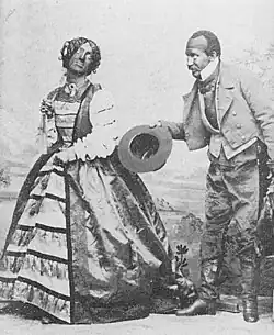 Two blackface performers on stage, one a man dressed in fancy woman's clothes, the other in dress attire, bowing with his hat in his hand.