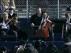 Four male musicians playing a variety of instruments outdoors in front of a chain-link fence