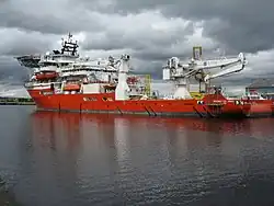 A red hulled ship seen from the port quarter at a low angle. It has heavy lifting gear on the quarterdeck and a helipad over the forecastle.