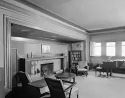 The living room's north and east walls. The north wall to the left has a fireplace, and the east wall to the right has windows. There is furniture arranged around the room.